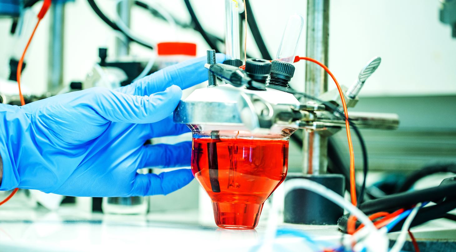 Blue lab glove holding a glass filled with red liquid in an electrochemical setup